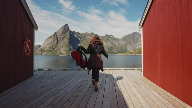 red head girl runs on the pier towards the sea next to the traditional red wooden rorbu fisherman's house on the seashore in the Lofoten archipelago, northern norway