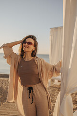Happy fair-skinned young model girl posing at camera holding her head on beach. Brown-haired woman with straight hair and closed eyes wears sunglasses. Rest time concept.