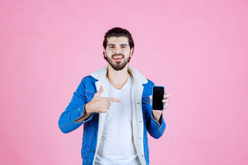 Young person holding a smartphone and making a peace sign gesture against a pink background, dressed in a blue jacket and white shirt, capturing a casual and cheerful vibe