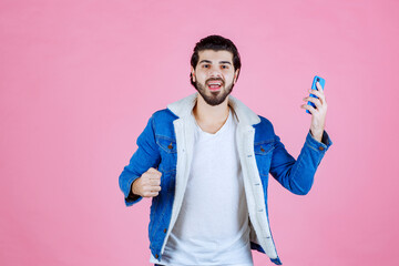 Young boy holding a smartphone and making a peace sign against a pink background, wearing a blue jacket and casual attire, showcasing confidence and modern technology usage