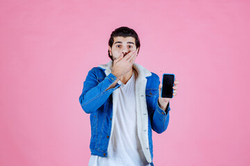 Young man with a backpack and smartphone standing against a pink background, dressed casually in a blue jacket and white shirt, capturing a selfie or taking a photo with his phone