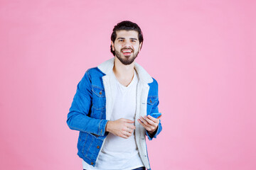 Young man in casual attire holding a smartphone against a pink background, wearing a blue jacket and white t-shirt, capturing a candid moment