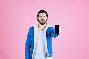 Young man taking a selfie with a smartphone in front of a pink background, wearing a blue jacket and white hoodie, capturing a casual and vibrant moment