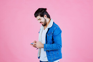 Young woman in a blue denim jacket holding a smartphone against a pink background, casual fashion, modern lifestyle, focused on technology use