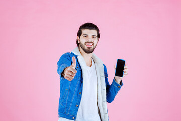 Young person holding a smartphone and giving a thumbs-up gesture against a pink background, wearing a blue jacket and white shirt, conveying positivity and technology engagement