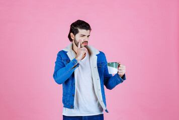 Young man with a blurred face wearing a blue jacket and white hoodie, holding a smartphone and a beverage against a solid pink background