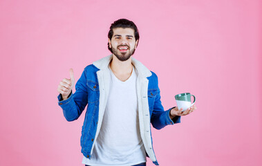 A person standing against a pink background holding a metallic bowl, wearing a blue jacket over a white shirt, with a blurred face for anonymity