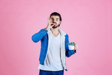 Young man in casual attire holding a beverage and a smartphone against a solid pink background