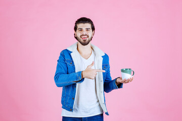 Young man with blurred face holding a small container and pointing at it, standing against a pink background, dressed casually in a blue jacket, white t-shirt, and glasses
