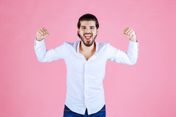 Person with short hair wearing a white shirt and blue jeans, posing with flexed arms in front of a pink background, showcasing strength and confidence