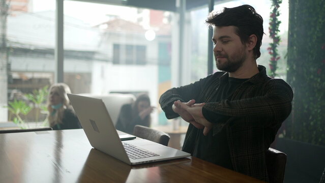 Young Man Stretching Body In Front Of Laptop Sitting At Coffee Shop. Person Stretches Arm Staring At Computer Screen