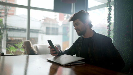 Young man looking at cellphone inside coffee shop in front of laptop. Person browsing internet on smartphone at cafe place