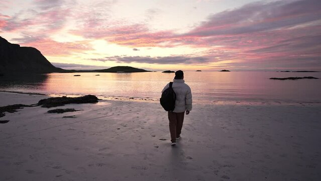 positive guy with a backpack looks at the sunset on the sandy beach. norway lofoten