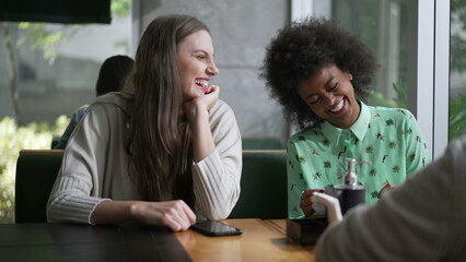 Three young diverse women laughing and smiling sitting at coffee shop table. Interaction between girlfriends in conversation. Real life laugh and smile - obrazy, fototapety, plakaty