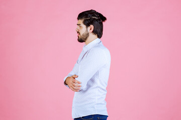 A young man with dark hair and a beard, wearing a light blue button-up shirt, stands with arms crossed against a solid pink background, creating a vibrant and modern portrait scene