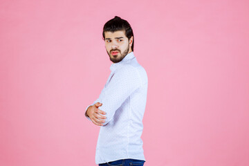 Young man standing with arms crossed against a pink background, wearing a white shirt and dark jeans, with a confident posture and casual style.