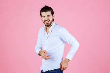 A man in a white shirt standing against a pink background, posing with one hand on his hip and the other holding an object, exuding confidence and style.