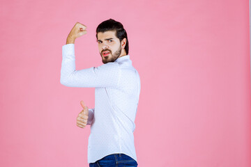 A woman standing against a pink background, flexing her right arm to show strength and confidence. She is dressed in a white top and dark jeans, with her hair tied back.