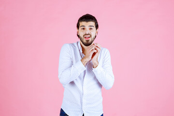 Person standing against a pink background holding a smartphone, dressed in a white shirt with long sleeves and dark pants, capturing a photo or selfie with a focused pose