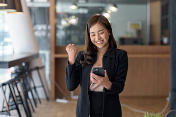 Beautiful Asian businesswoman using smartphone is delighted and excited by the success she has achieved through her office cell phone.