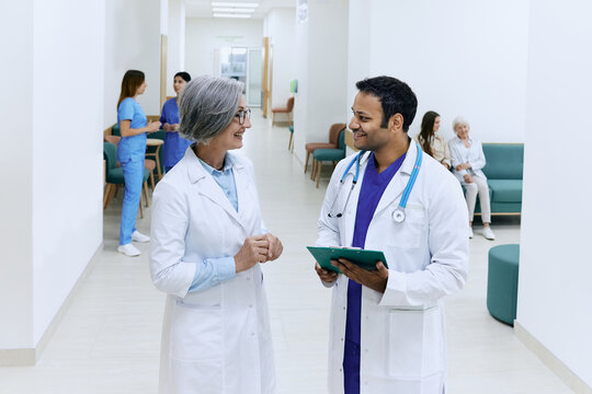 Council Of Doctors. Mature Female General Practitioner Communicates With Indian Doctor While Working Day Standing In Hospital