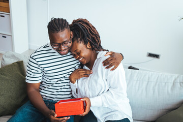 Fototapeta premium Shot of a loving husband giving his wife a gift. Boyfriend surprise his beautiful girlfriend with present while she is sitting on the sofa in the living room at home. Romantic couple with present.