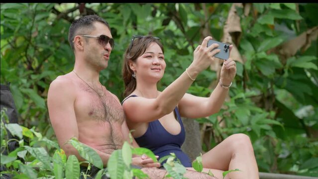 A couple making selfie picture using cellphone as they are sitting in the jungle forest at honeymoon vacation. Girl making faces and smiling to boyfriend as they are posing for photographs in woods