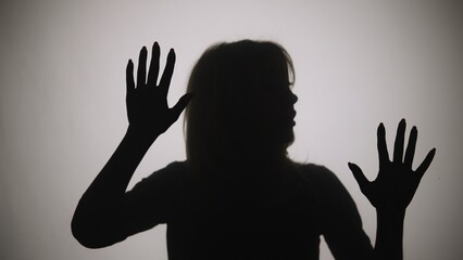 Black silhouette of a woman touching the glass with her palms and looking at the camera. The outline of a woman against the background of a window with white matte light. Close up.