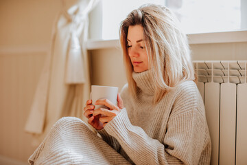 Young woman in long winter beige sweater is posing at home near the radiator