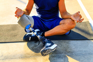 man with prosthesis leg practicing yoga sitting in lotus pose on yoga mat meditating outdoors in...