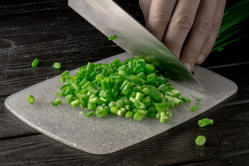 Male hand cutting green onion stalks with knife into small slices. Metal knife chopping shallots or chive on gray plastic board on wooden restaurant kitchen table. Chef cooking vegan salad. Close up.