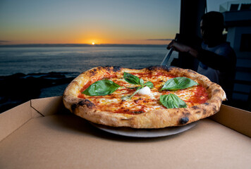 Pizza al estilo italiano con diferentes ingredientes con el fondo de un atardecer en el que se ve el sol y el mar - obrazy, fototapety, plakaty