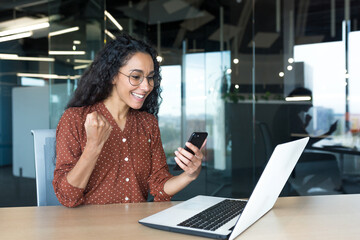 Young successful hispanic businesswoman working inside modern office, female worker using laptop, holding phone smiling and happy celebrating victory and triumph.