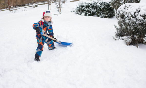 5-6 year old boy in bright warm overalls, helps to clear the yard with snow after a snowstorm, throwing snow away with a big shovel. Winter season, bad weather, cold, snowy