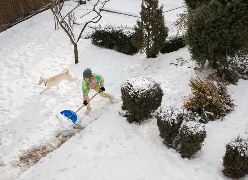 young woman in bright warm sportswear, clearing sidewalk, street, yard from snow after a strong snowstorm, throws snow on shovel to side. Winter season, bad weather, cold, snow. dog running around