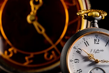 Silver antique swiss pocket watch with gold hands and vintage table clock in blurry background. An old round white dial of pocket watch and brown face of table clock with hands. Close up.