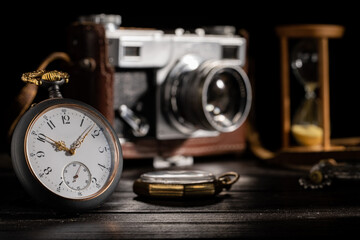 Antique gray pocket watch with a retro film camera and hourglass on blurred background. Silver round pocket watch with golden hands, camera in brown leather case on dark wooden table. Close up.