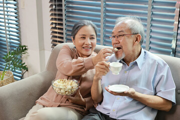Happy smiling asian senior man and woman sitting on sofa and eating popcorn while having fun with movie rest indoor at home living room. Couple elder husband and wife embrace are happy while watch TV