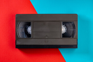 Black vintage VHS videotape cassette on red and blue background. Plastic retro video cassette with analog magnetic tape. Old technology for tape recording and watching media movies. Top view. - obrazy, fototapety, plakaty