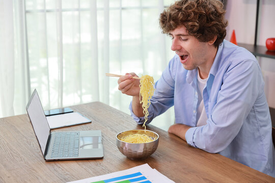 Young Caucasian Currly Hair Man Sitting On Table And Eating Tasty And Instant Noodle For Lunch In The Working Room With Chopsticks And Computer. Unhealthy And Cheap Food Lifestyle Conceptncept.