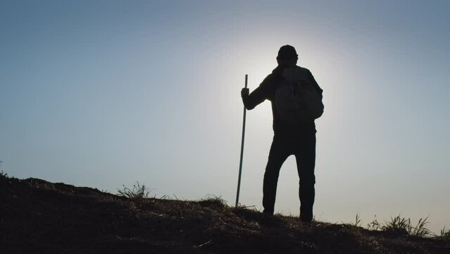Adventurous man stands on high hill spreading hands to sides with joy at back light after throwing trekking pole and backpack on ground. Silhouette of traveler against blue sky at sunset backside view