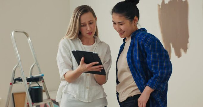 A blonde woman, holding a tablet in her hands, gives her friend recommendations for effective home repairs. The two are chatting and laughing. They like what they are doing now.