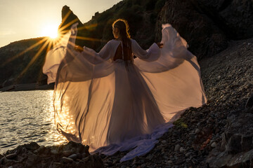 A mysterious female silhouette with long braids stands on the sea beach with mountain views, Sunset rays shine on a woman. Throws up a long white dress, a divine sunset.