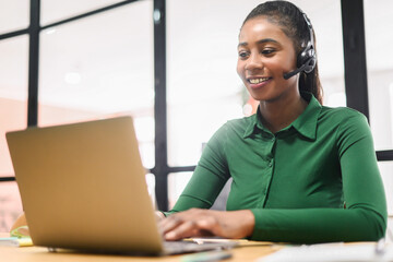 Smiling african-american female employee colleague using headset and laptop sitting on the workplace, talking on the distance, work in call center, multiracial woman is customer support operator