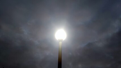 A street lamppost illuminates the neighborhood. Under a dark, gloomy sky covered with clouds, there is a long iron lamppost with a lamp on top. The lantern shines brightly in the dark.