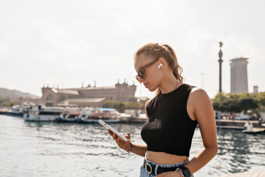 Profile outside photo of stylish european woman with dark hair wearing summer clothes using smartphone and listening music on background of lake with yachts in sunny warm day