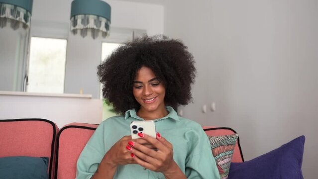 Calm and carefree african lady sitting in a relaxed pose on the comfortable couch with a smartphone, woman spends leisure time scrolling news feed, web surfing, texting with friends