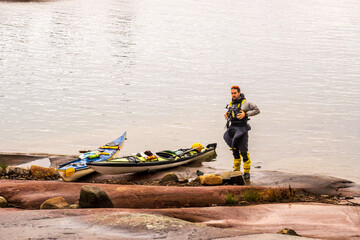 Obraz premium A sea kayaker in safety gear stands beside his boat on the rocky shore of Georgian Bay in the evening light. Room for text.