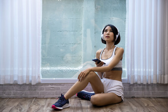 Asian woman in sportswear listening to music with headphone connected via smartphone after exercise at home fitness.