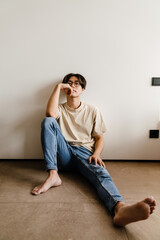 Asian young man thinking while sitting on floor by white wall at home